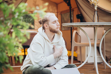 Young man works on laptop while sitting relaxed with cup of coffee in camp tent.