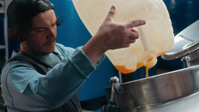 Chest up of Caucasian male brewmaster in backward baseball cap filling stainless steel tank with orange liquid in plastic canister