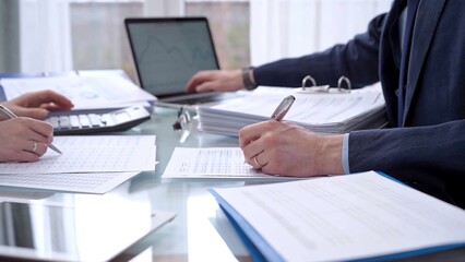 Business people wearing blue suits are reviewing financial data in office. Two professionals analyzing paper reports using calculators. Audit and taxes concept