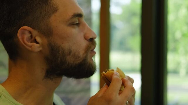 A young bearded man is sitting in a cafe and biting a delicious fresh hot meat burger with cheese. The guy is sitting at a table by the window in a cafe and enjoying a break alone with cheeseburger