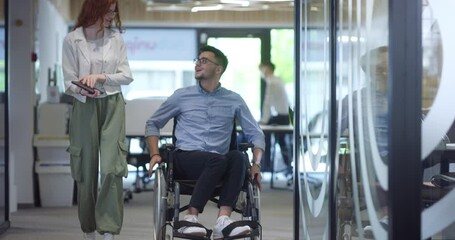 Young business colleagues, collaborative business colleagues, including a person in a wheelchair, walk past a modern glass office corridor, illustrating diversity, teamwork and empowerment in the