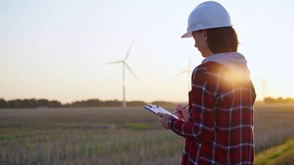 Adult woman engineer wearing white cask is taking notes on a clipboard on a field with wind turbines, as the sun sets, back view