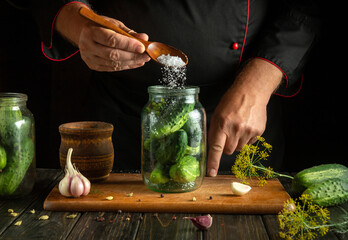 An experienced cook adds salt to a jar of cucumbers with a spoon. The concept of pickling cucumbers on the kitchen table.