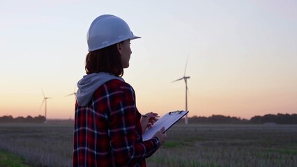 Woman engineer is taking notes on a clipboard on a field with wind turbines, as the sun sets in evening. Clean energy and engineering