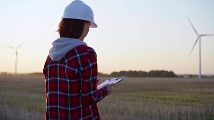 Adult woman engineer wearing white cask is taking notes on a clipboard on a field with wind turbines, as the sun sets, back view