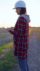 Woman engineer wearing a white protective helmet is taking notes with a clipboard in a field with wind turbines. Clean energy and engineering concept