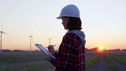 Woman engineer is taking notes on a clipboard on a field with wind turbines, as the sun sets in evening. Clean energy and engineering