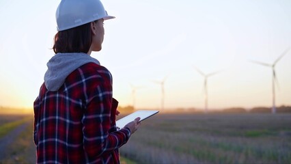 Female engineer taking notes with a tablet computer on a field with wind turbines, as the sun sets....