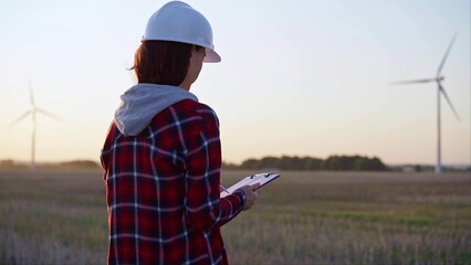 Adult woman engineer wearing white cask is taking notes on a clipboard on a field with wind turbines