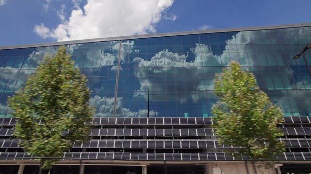 Solar panels and affordable renewable energy source in layers and rows are lined on the side of a large business and glass high rise siding and wall as the sun hits the upper class building