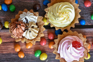Colorful tartlets cakes with protein cream, multicolored dragee candies on wooden table. Delicious dessert with chocolate, cherry and banana flavors, shortcrust pastry. Party food. Top view.