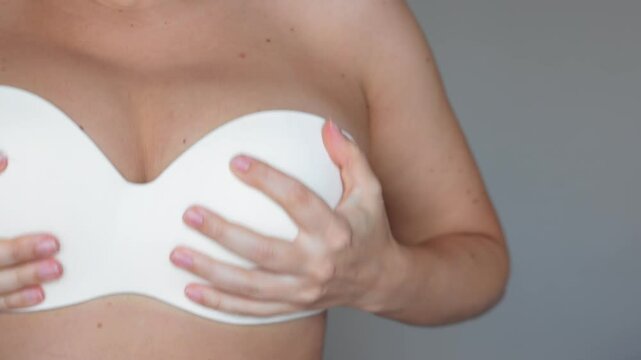 Close up of a a young woman pulling her bra up lifting her saggy breasts on a grey background. The result of breastfeeding