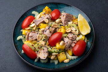 Salad with canned tuna, tomatoes, peanuts, mango, avocado, celery, chili flakes and black sesame. Light summer salad with olive oil and a lemon lobule. Selective focus, close-up, black background.