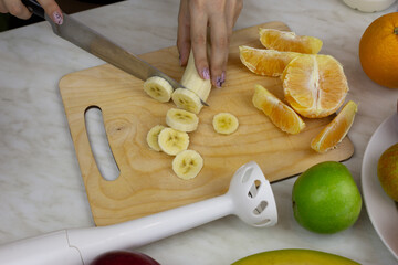 Female hands cutting banana on cutting board in kitchen. Making fruit drink in blender for health and sport. Fruit smoothie ingredients on table