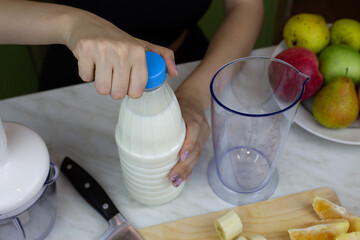 Girl opens bottle of milk to use in blender with fruits while making milkshake. Sports drink with vitamins from fruits and milk, ingredients for healthy drink recipe