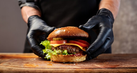 A close-up of a fresh burger being prepared by black-gloved hands. The burger features a juicy patty, fresh lettuce, tomato slices and melted cheese, all coming together on a bun.