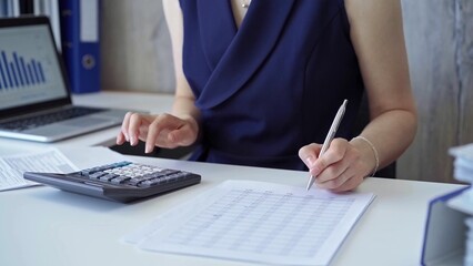 Businesswoman with dark blue dress is using dark calculator and taking notes of company expenses in her office. Taxes and audit in business