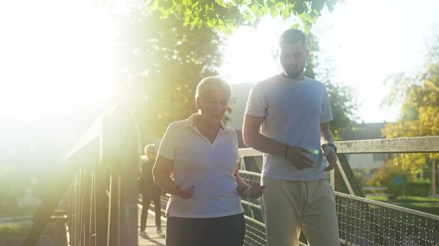 Mother and Son Crossing Pedestrian Bridge at Sunset. Physiotherapist Consulting Client in Nature. Senior Woman and Young Man Strolling Together. Concept of explaining, rehabilitation, mentor, advice
