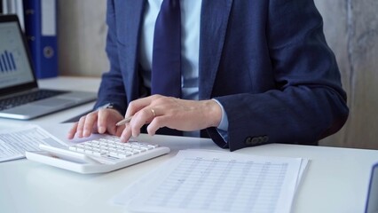 Businessman accountant with blue suit is using white calculator and taking notes with a silver pen. Taxes and audit concept