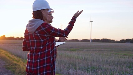 Man and woman, both engineers wearing white protective helmets, are checking and discussing something in a field with wind turbines as the sun sets. Clean energy and engineering concept