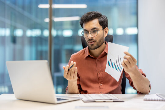 Businessman in office holding paper chart, explaining financial data during video call on laptop. Focus on analysis, communication, technology. Reflects professional environment and office work - Powered by Adobe
