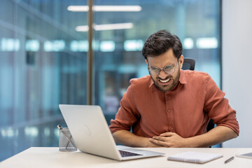 Business professional in office showing signs of abdominal pain and nausea while using laptop. Man wearing glasses, appearing stressed and uncomfortable, holding stomach.