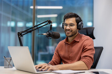 Smiling man wearing headphones recording audio podcast with professional microphone setup and laptop in modern office. Engaged in work, representing creativity, technology, and media production