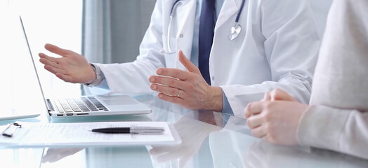 Doctor is consulting patient in modern fair clinic. Healthcare professional discusses medical information with a woman, gesturing above the laptop computer on the glass desk. Medicine