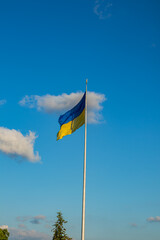 Ukrainian flag waving on a tall flagpole against a blue sky with scattered clouds on a sunny day.
