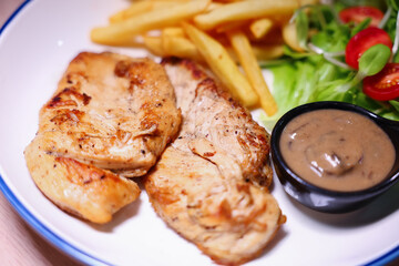 Chicken steak with side dishes of French fries, salad and pepper sauce on a white plate, photographed with studio lights.