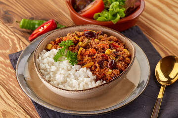 A hearty bowl of chili con carne with white rice, featuring ground beef, beans, corn, and peppers, garnished with parsley. Served alongside a fresh salad on a rustic wooden table.
