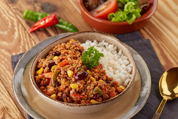 A hearty bowl of chili con carne with white rice, featuring ground beef, beans, corn, and peppers, garnished with parsley. Served alongside a fresh salad on a rustic wooden table.