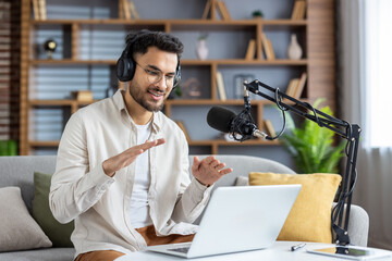 Confident young man recording podcast at home while using laptop and microphone, engaging with...