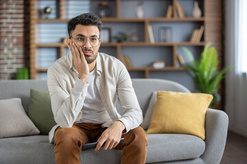Bored man sitting on sofa with remote control, feeling disinterested while watching television in modern living room at home.