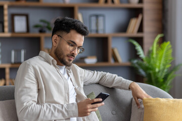 Relaxed man sitting on sofa using smartphone, browsing internet or checking social media in cozy living room with modern decor