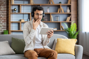Man on sofa wearing headphones, surprised while watching something on tablet in cozy living room