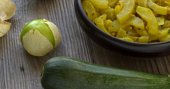 Homemade zucchini curry with tomatillo, garlic, onion and spices. On the side are shown the main ingredients: zucchini, tomatillo, and garlic bulb. Table spin.