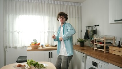 Happy father move to music while cooking food and prepare lunch at modern kitchen. Active dad dancing to song while making healthy food or salad surrounded with vegetable and kitchenware. Pedagogy.