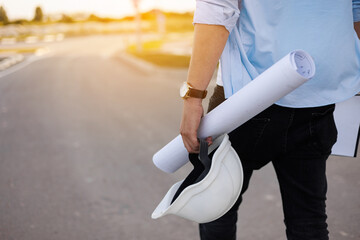 Unrecognisable engineer, architect or worker holds in hand protective white hard hat and blueprint on multi-apartment residential complex background, sunlighting. Concept of new real estate.