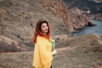 Portrait of a happy woman with long hair against a background of mountains and sea. Holding a bouquet of yellow tulips in her hands, wearing a yellow sweater