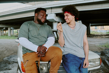 Biracial best friends smiling to each other while curly haired one smoking cigarette, they leaning...
