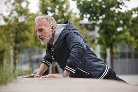 Mature fit man doing push ups on bench working out regularly in city park, copy space