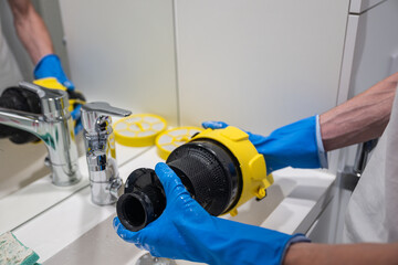 Hands in blue gloves washing 
vacuum cleaner filter with water in bathroom sink