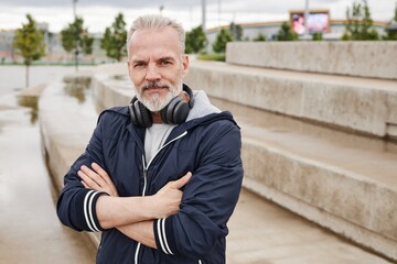 Fototapeta premium Medium portrait shot of smiling senior man in navy windbreaker on morning workout at city park posing with folded arms and looking at camera, copy space