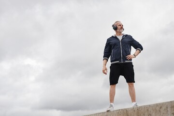 Full shot of pensive senior man in navy windbreaker and headphones on morning workout at city park posing with arm on waist and looking away against gray overcasted sky, copy space