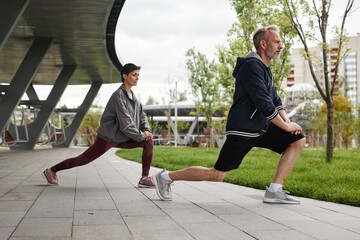 Side view of fit senior man and woman in static lunge pose strengthening leg muscles, while working out together under shade pavilion at autumn city park