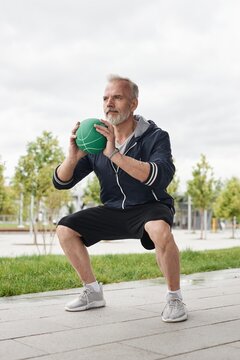 Vertical shot of fit senior man holding ball in static squat position working out at city park against gray autumn sky, copy space