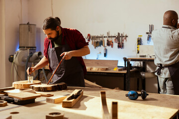Craftsperson in carpentry shop starting restoration project on wood block, applying lacquer on surface next to coworker in background. Carpenter coloring plank with paintbrush near BIPOC colleague