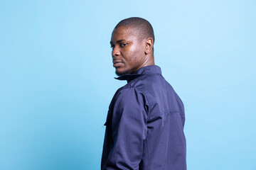 Portrait of african american security agent posing in front of camera, looking over his shoulder against blue background. Male bodyguard dressed in uniform, detecting any suspicious behavior.