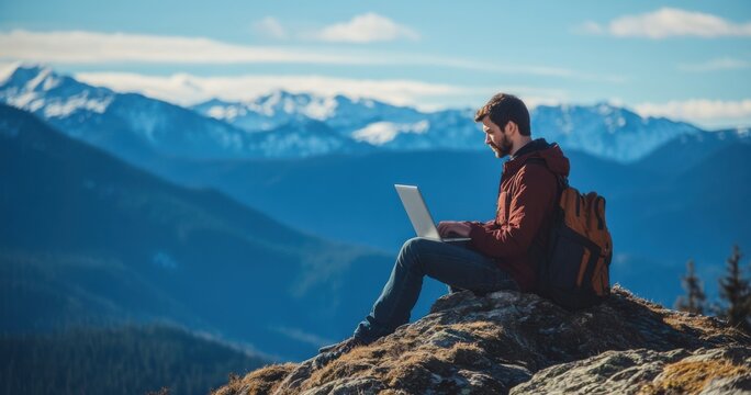 A young man works on a laptop while sitting at the top of a mountain. This concept represents the digital nomad lifestyle and the ability to work remotely or travel using the internet.
